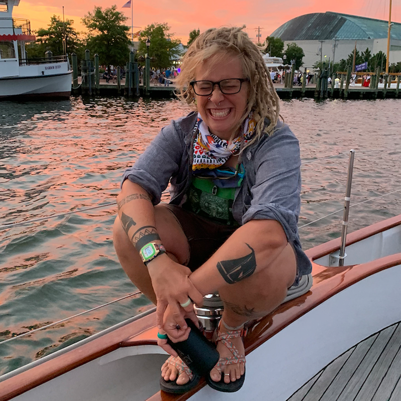 The image shows a woman with dreadlocks, glasses, and tattoos, sitting on a boat. She is smiling and appears to be enjoying herself. The background features a sunset over the water, with other boats and a building visible. The overall scene suggests a relaxed and happy atmosphere.